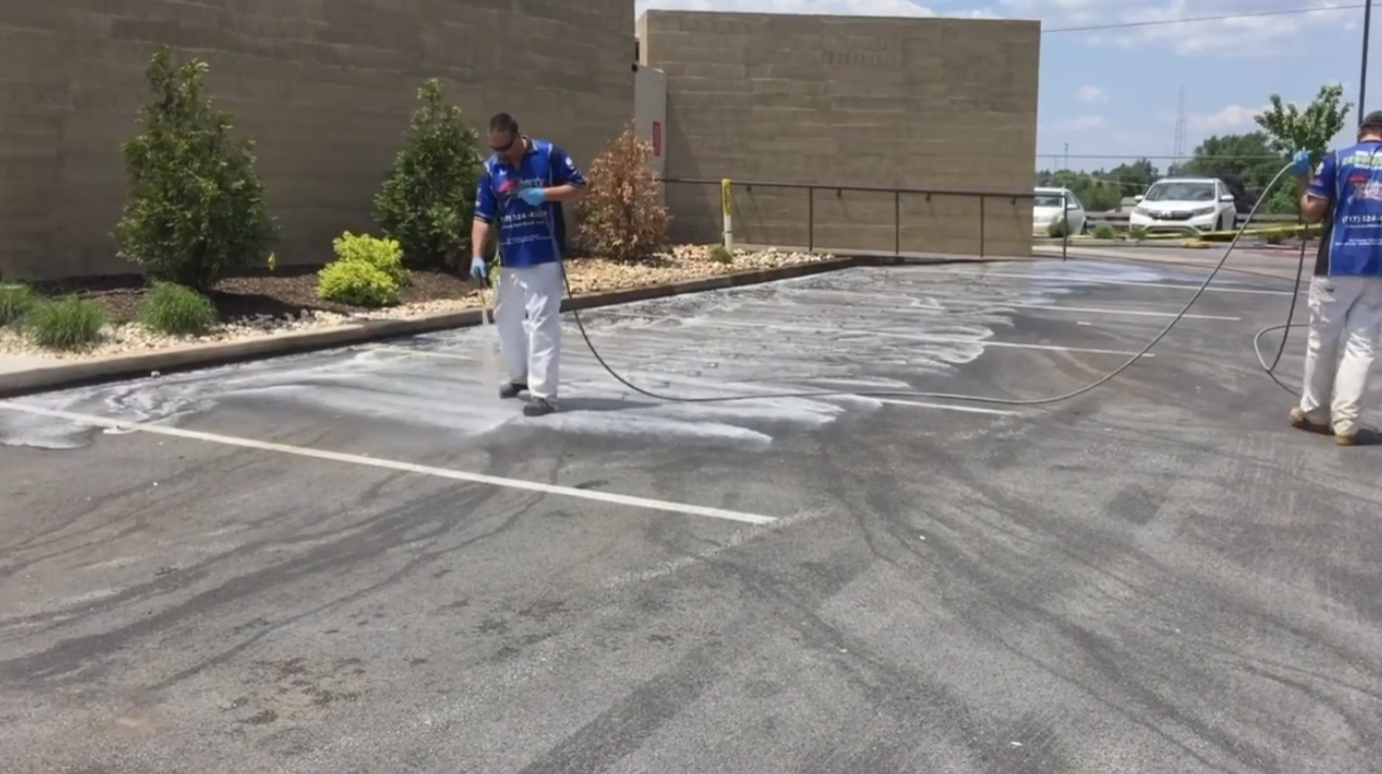 Two workers power-washing a parking lot; soapy water spreads across the asphalt as they spray with hoses outside a building.