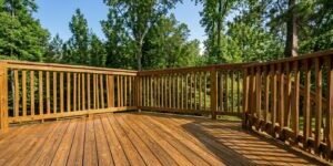 Sunlit wooden deck with railing overlooking a dense green forest under a blue sky and tall trees.