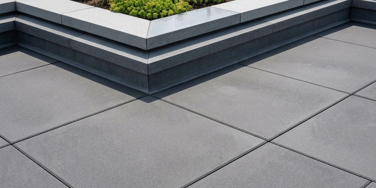Raised rectangular planter on a flat grey concrete-tiled terrace with green shrubs and grasses along the edge; multi-level concrete edging creates a small garden bed.