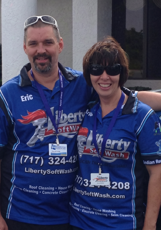 Two smiling people in matching blue Liberty SoftWash uniforms pose arm in arm outdoors with badges around their necks.