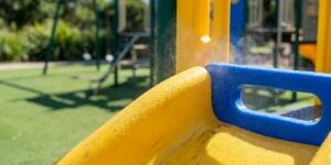 Close-up of a yellow playground slide with a blue handrail, water droplets on the surface and a blurred grassy playground in the background.