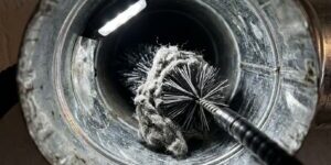 Inside a metal duct/vent, a round stiff-bristle brush on a long wand is used for cleaning. A rag is wrapped around the brush.
